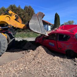 Ein gelbes Baufahrzeug hebt Holzschnitzel über ein beschädigtes rotes Auto auf einer Baustelle.