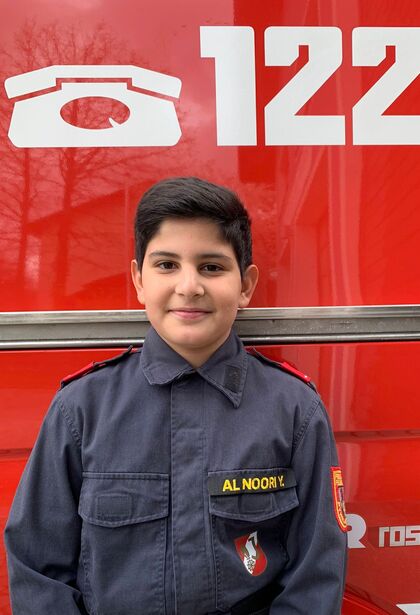 Ein lächelnder Junge in Feuerwehruniform steht vor einem roten Feuerwehrfahrzeug mit der Nummer 122.