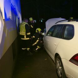 Feuerwehrleute inspizieren bei Nacht ein weißes Auto mit geöffnetem Kofferraum und Motorhaube nahe einer Gebäudewand.