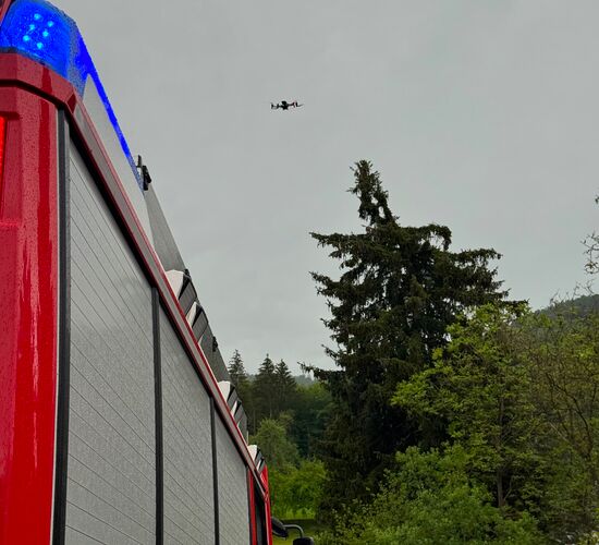 Ein Feuerwehrfahrzeug mit Blaulicht steht im Vordergrund, während eine Drohne am Himmel fliegt. | © Michael Schuster