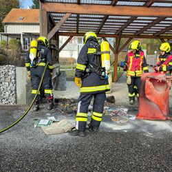 Feuerwehrleute inspizieren eine verbrannte Mülltonne unter einem Carport neben einem Wohnhaus.