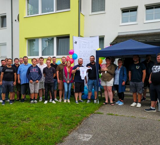 Foto: Alle Gratulanten vor dem aufgestellten Storch, Kommandant und Papa halten einen der überreichten Bodys für die Kleine Annika