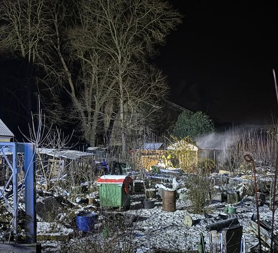 Ein winterlicher Garten bei Nacht, zeigt Bäume und einen Schuppen, aus dem Rauch aufsteigt unter einem klaren Himmel.