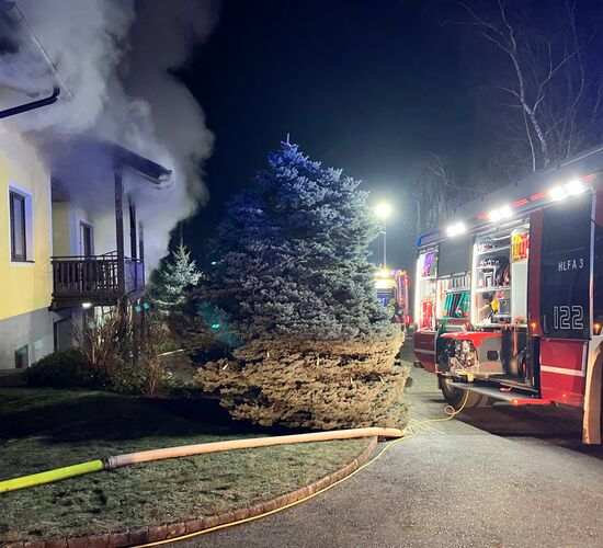 Ein Feuerwehrauto steht vor einem rauchenden Haus, während Löschschläuche im nächtlichen Einsatz bereitliegen.