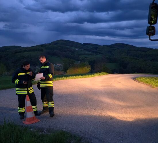 Zwei Feuerwehrleute sprechen über Funk neben einem orangefarbenen Warnkegel auf einer Straße in der Abenddämmerung.