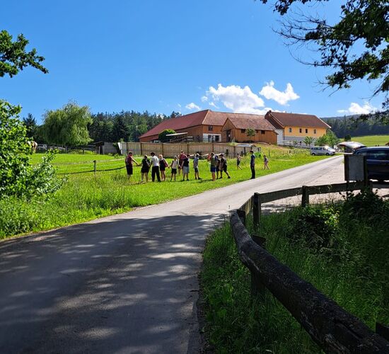 Eine Gruppe von Menschen versammelt sich auf einem Feld in der Nähe eines Bauernhauses an einem sonnigen Tag.