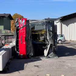 Ein umgekipptes rotes Fahrzeug mit zerbrochenem Glas steht auf einem Parkplatz, umgeben von Betonblöcken.