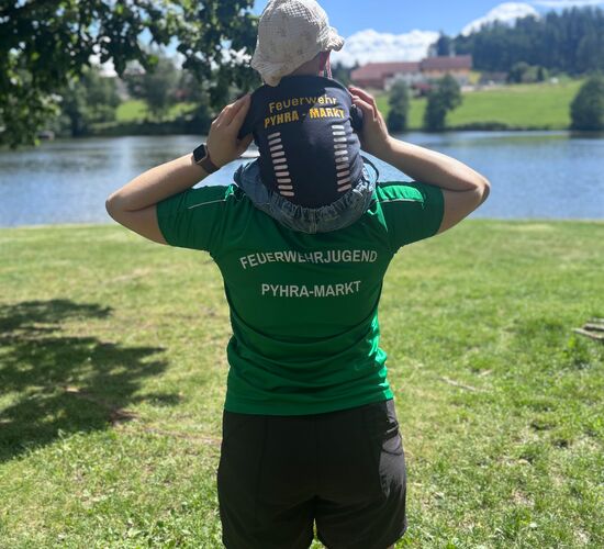 Ein Erwachsener mit einem Kind auf den Schultern steht am Wasser, beide tragen Feuerwehr-Pyhra-Marktkleidung.