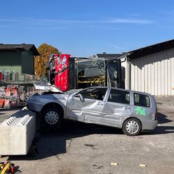 Ein beschädigter Pkw liegt auf einem Betonblock, während ein zerstörter Bus im Hintergrund sichtbar ist.