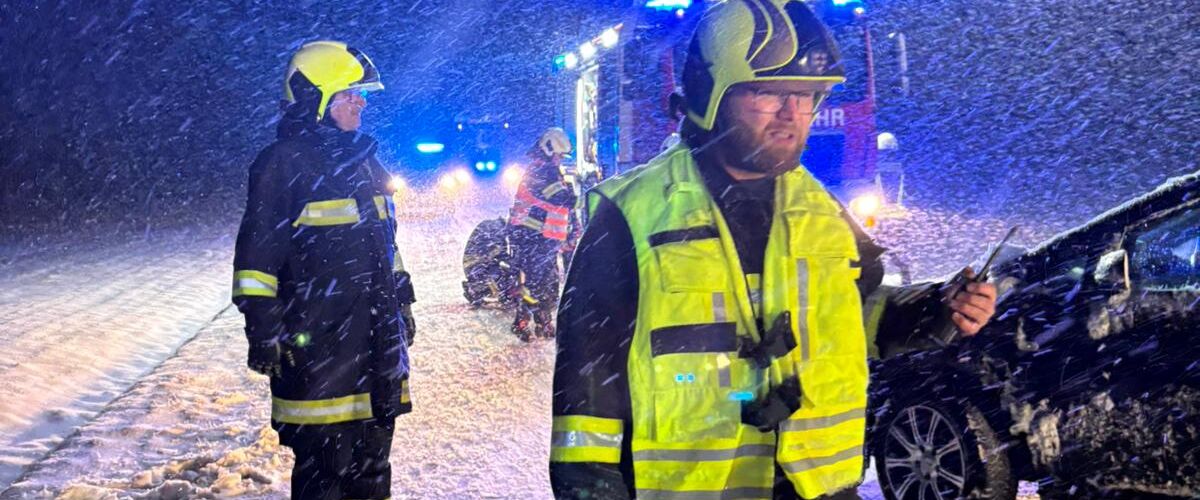 Feuerwehrleute arbeiten in einem Schneesturm bei Nacht auf einer verschneiten Straße, während ein Einsatzfahrzeug leuchtet.