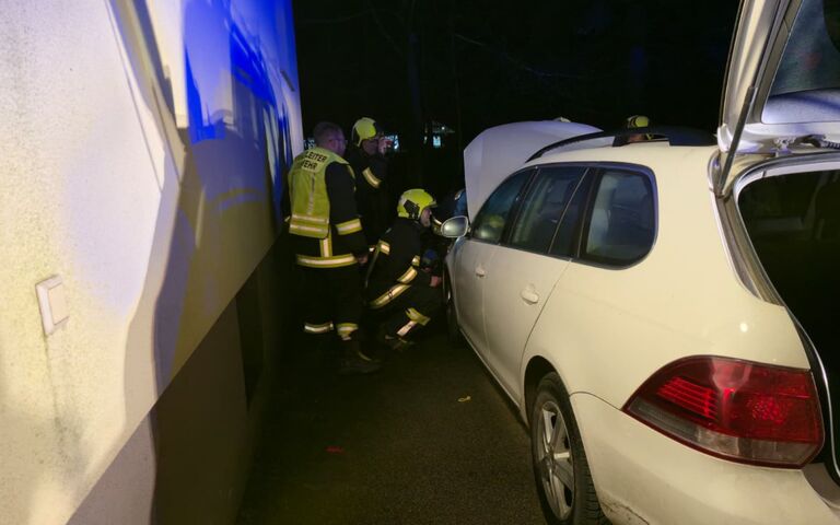 Feuerwehrleute inspizieren bei Nacht ein weißes Auto mit geöffnetem Kofferraum und Motorhaube nahe einer Gebäudewand.