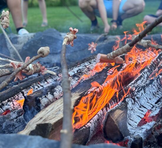 Ein Lagerfeuer brennt, während Personen in der Nähe Würstchen auf Stöcken rösten. | © Name