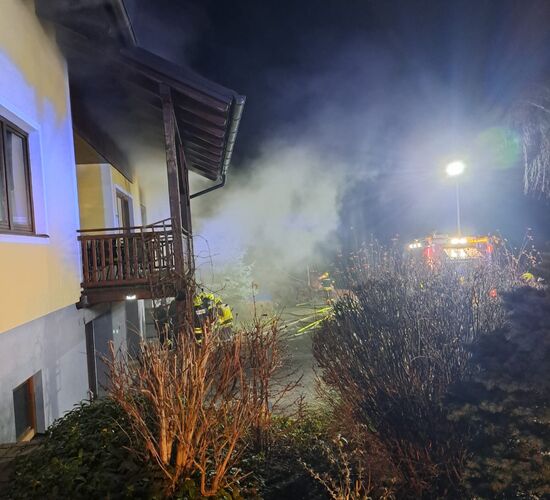 Ein nächtlicher Feuerwehreinsatz bei einem rauchenden Wohnhaus beleuchtet den Garten im Scheinwerferlicht.