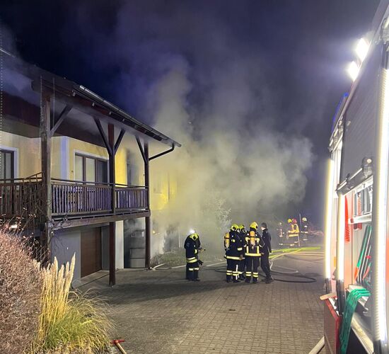 Feuerwehrleute bekämpfen einen nächtlichen Hausbrand, während dichter Rauch das Gebäude umhüllt.