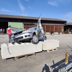 Ein silbernes Auto steht auf Betonblöcken, während Personen im Hintergrund beobachten und das Geschehen filmen.