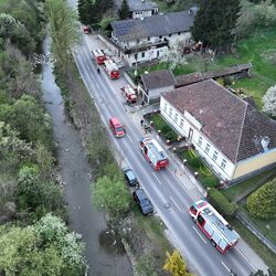 Luftaufnahme einer Straße mit Feuerwehrfahrzeugen und einem kleinen Fluss, umgeben von blühenden Bäumen und Häusern.