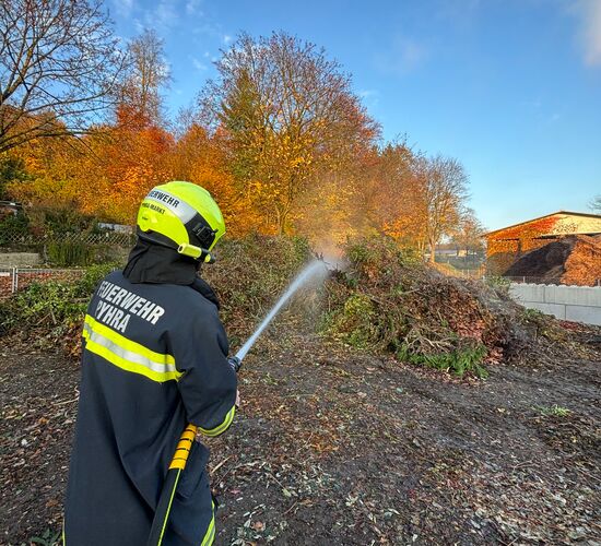 Ein Feuerwehrmann spritzt Wasser auf einen Grünschnitthaufen, während die bunten Blätter im Hintergrund leuchten.