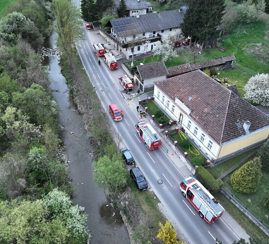 Luftaufnahme einer Straße mit Feuerwehrfahrzeugen und einem kleinen Fluss, umgeben von blühenden Bäumen und Häusern.