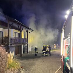 Feuerwehrleute bekämpfen einen nächtlichen Hausbrand, während dichter Rauch das Gebäude umhüllt.