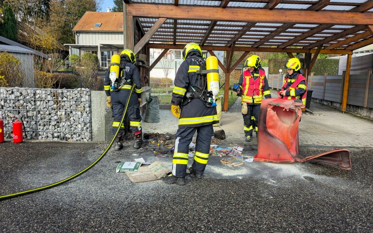 Feuerwehrleute inspizieren eine verbrannte Mülltonne unter einem Carport neben einem Wohnhaus.