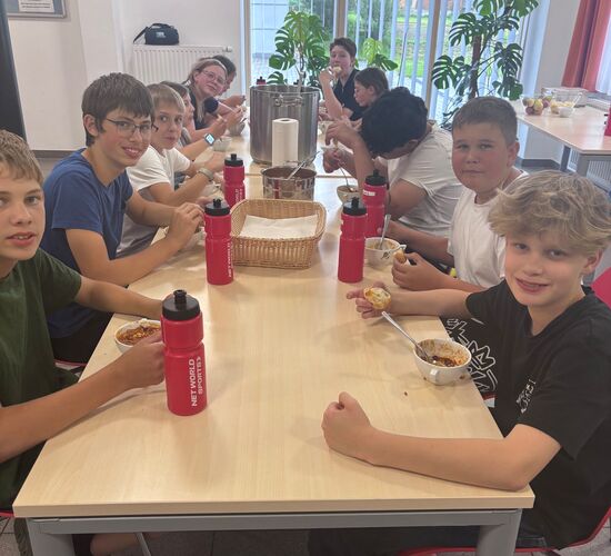 Eine Gruppe von Kindern genießt gemeinsam ihr Essen an einem Tisch, umgeben von Pflanzen und einem hellen Raum.