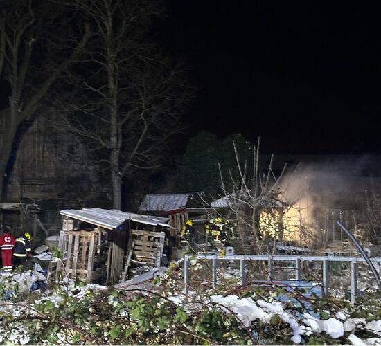 Bei Nacht löschen Feuerwehrleute ein brennendes Gartenhaus, während Helfer in roten Jacken Unterstützung leisten.